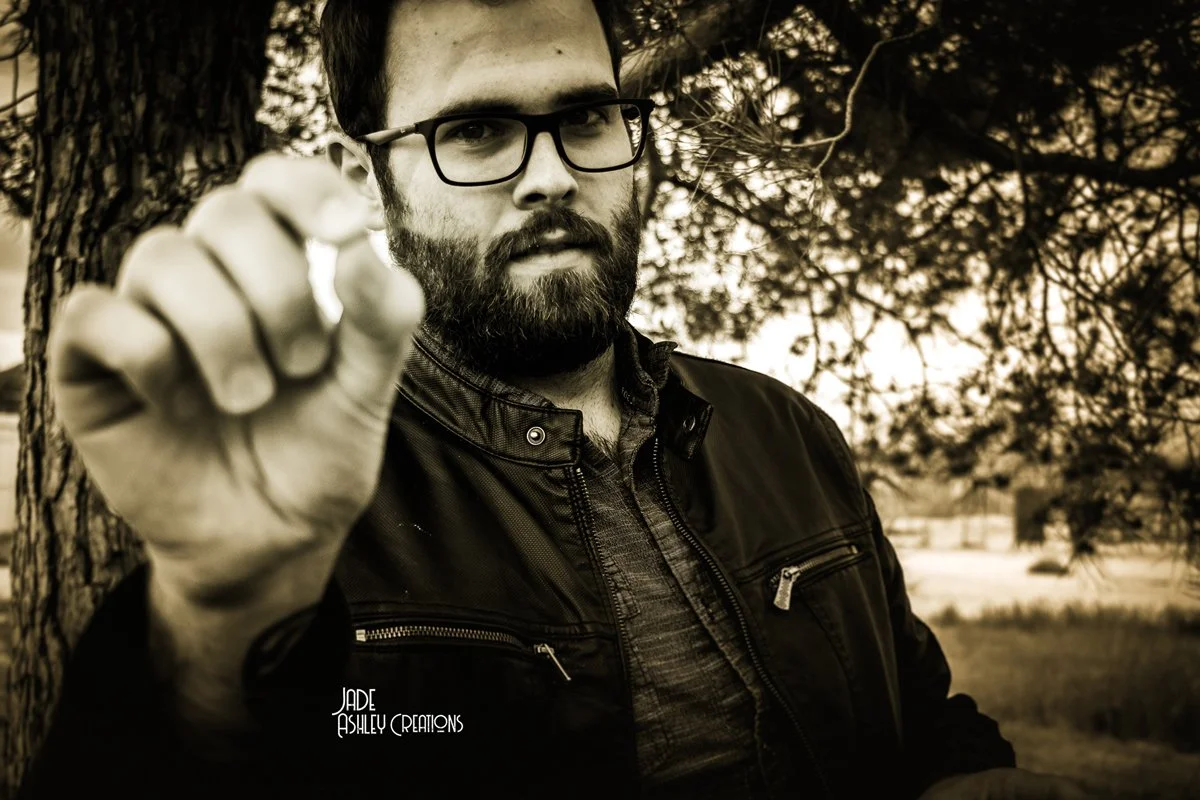 A man with glasses and a beard pointing towards the camera outdoors near a tree, black and white photo.