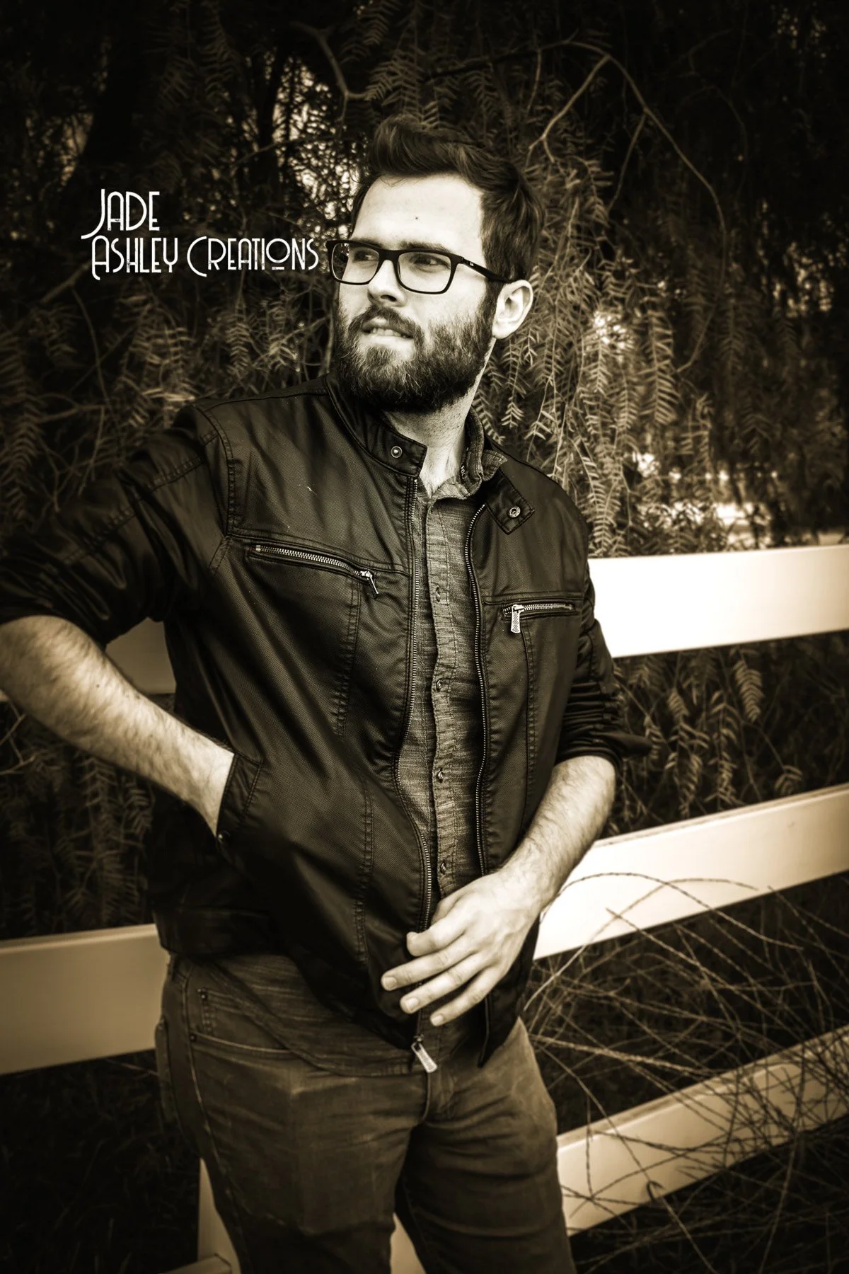 A bearded man wearing glasses, a dark leather jacket, and jeans, stands outdoors with a background of trees and branches, looking to his left with his right hand in his pocket and his left hand resting on his stomach.