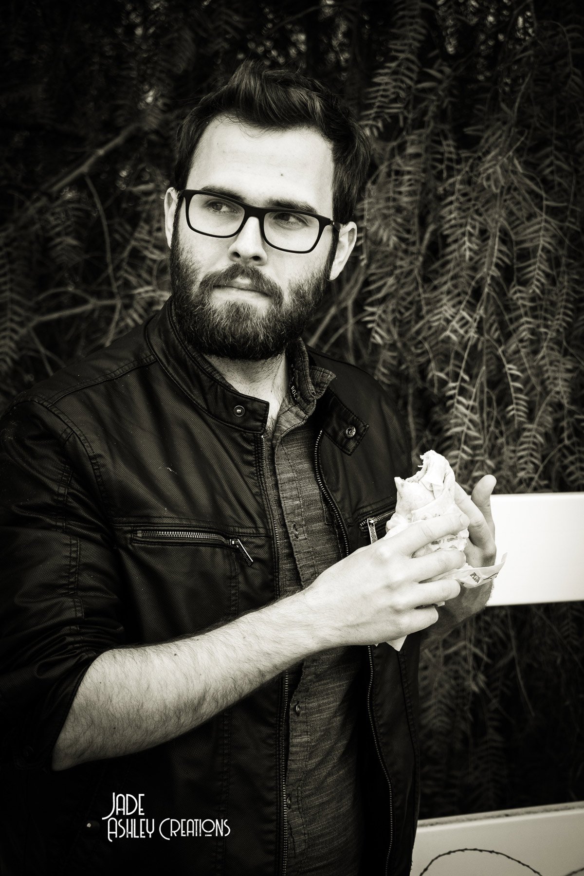 A man with dark hair, glasses, and a beard holding a wrapped sandwich. He is wearing a dark leather jacket and standing in front of leafy plants.