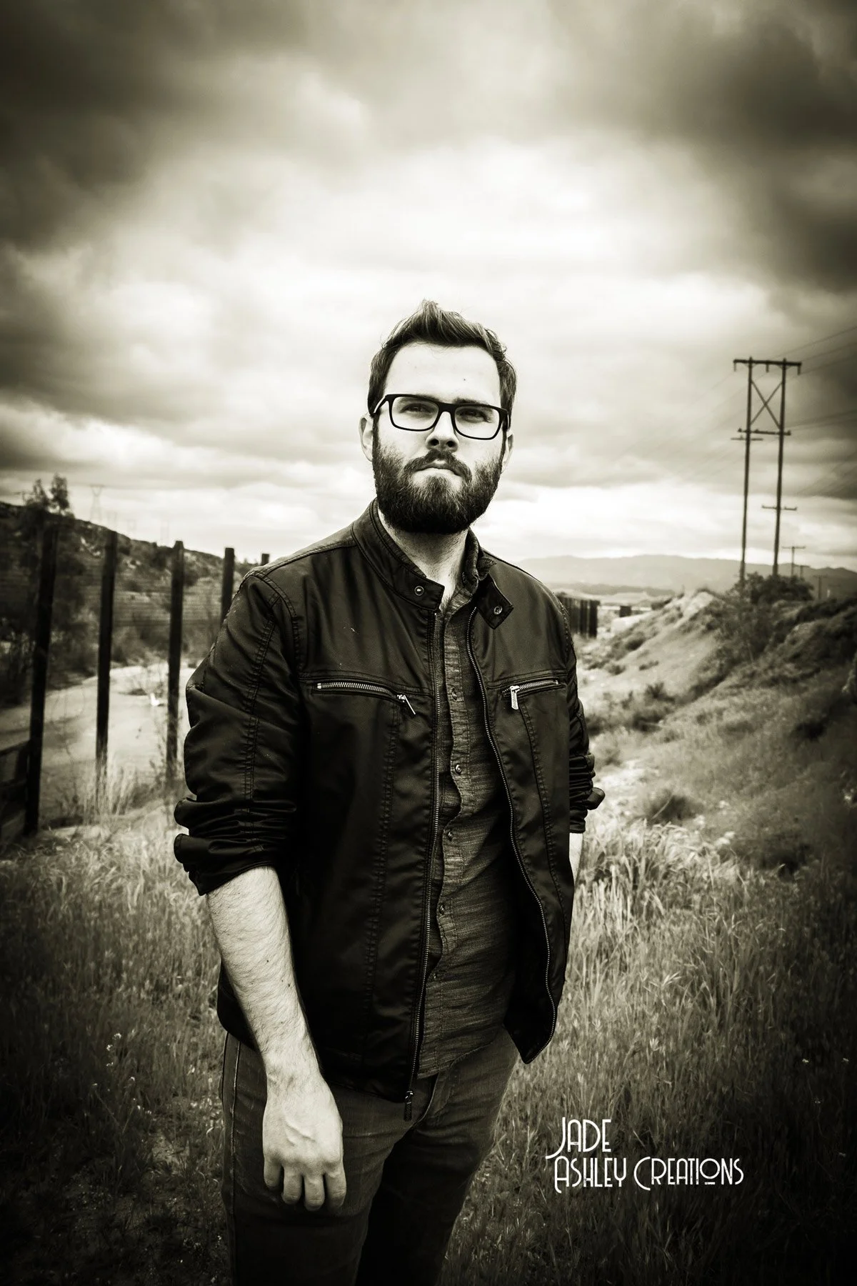 A black-and-white photo of a man with glasses and a beard standing outdoors in a rural area with grassy fields, a wooden fence, and power lines, under a cloudy sky.