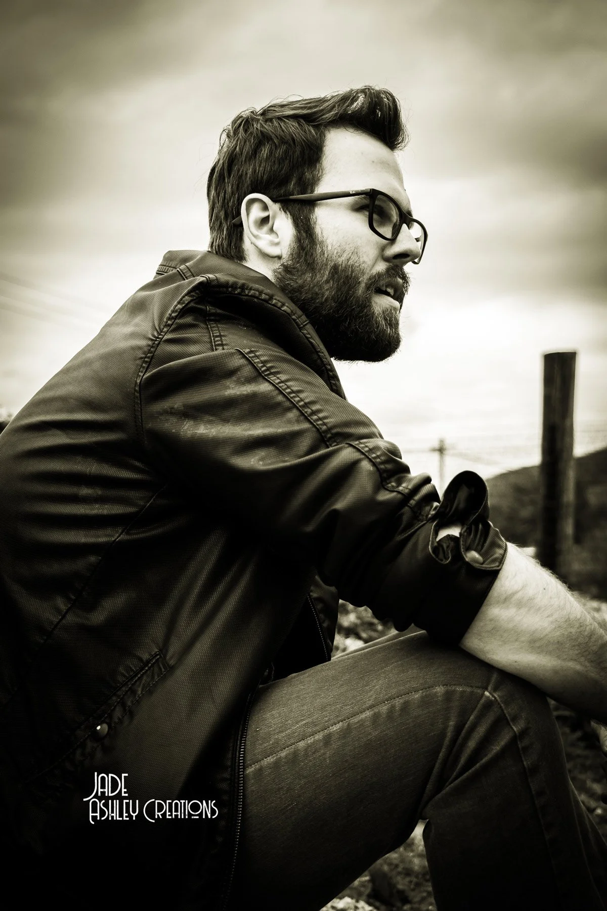 A black-and-white photograph of a man with glasses and a beard, sitting outdoors with a cloudy sky in the background, wearing a leather jacket and looking to the side.