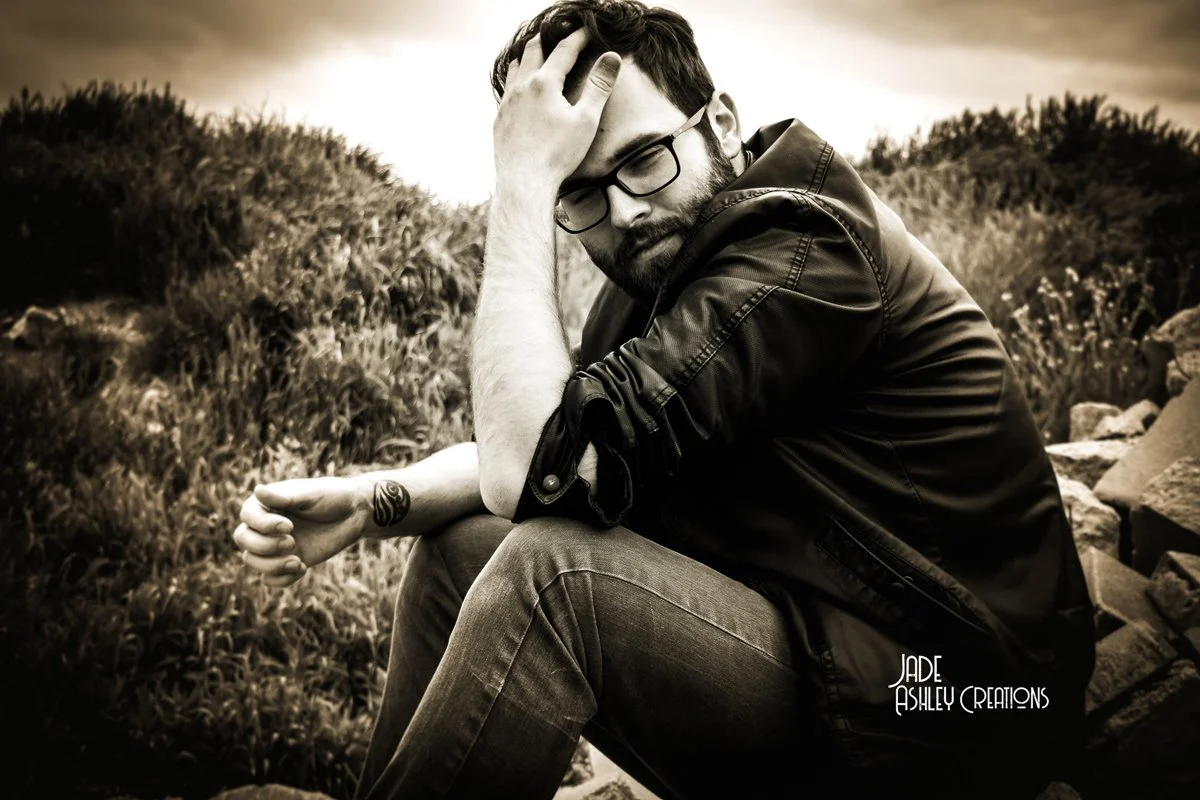 A man with glasses and a beard sitting outdoors on rocks, with one hand on his forehead and the other resting on his knee, appearing thoughtful or distressed.