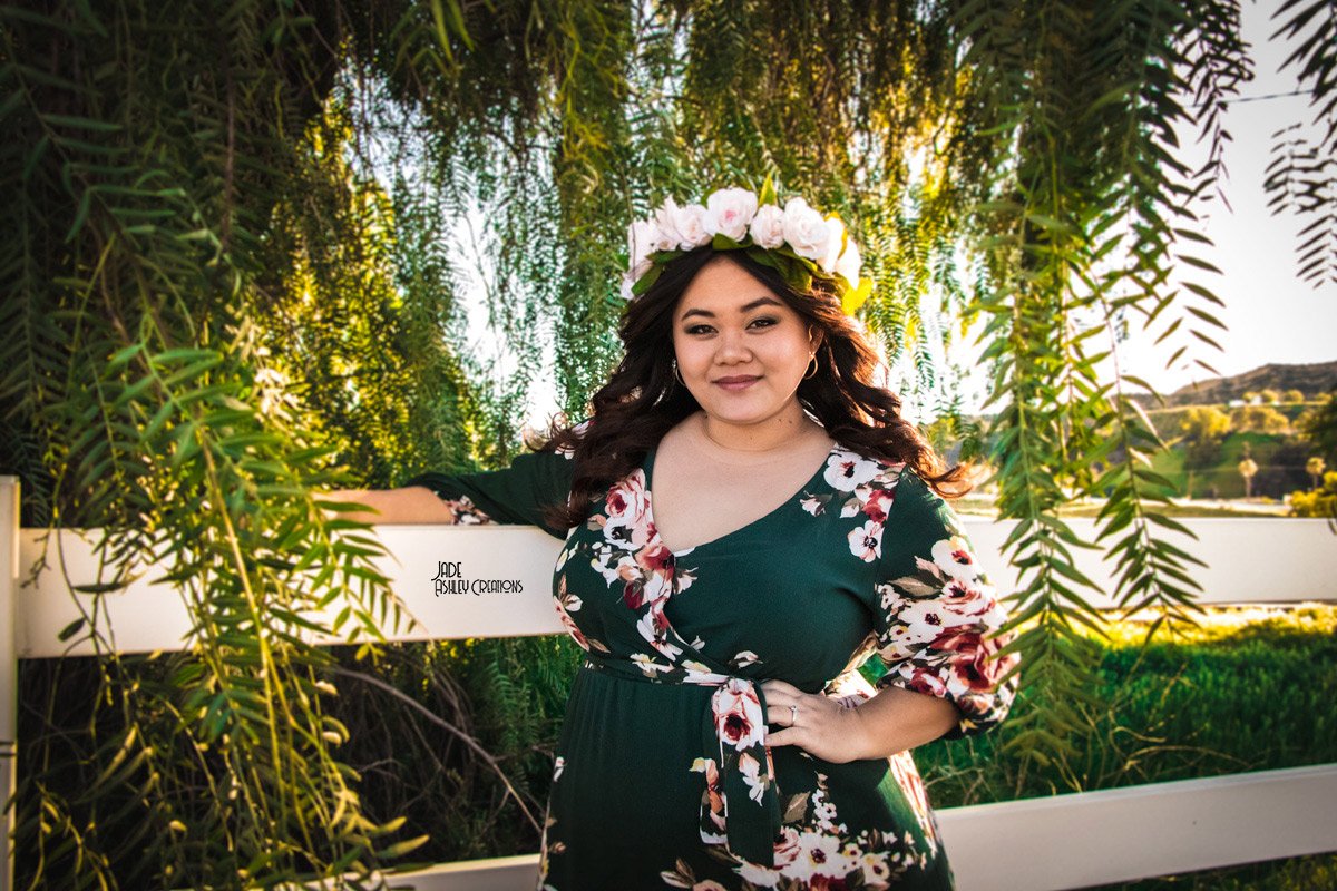 A woman with long dark hair and light skin, wearing a green floral dress with a matching floral crown, standing outdoors among hanging green plants and trees during sunset.