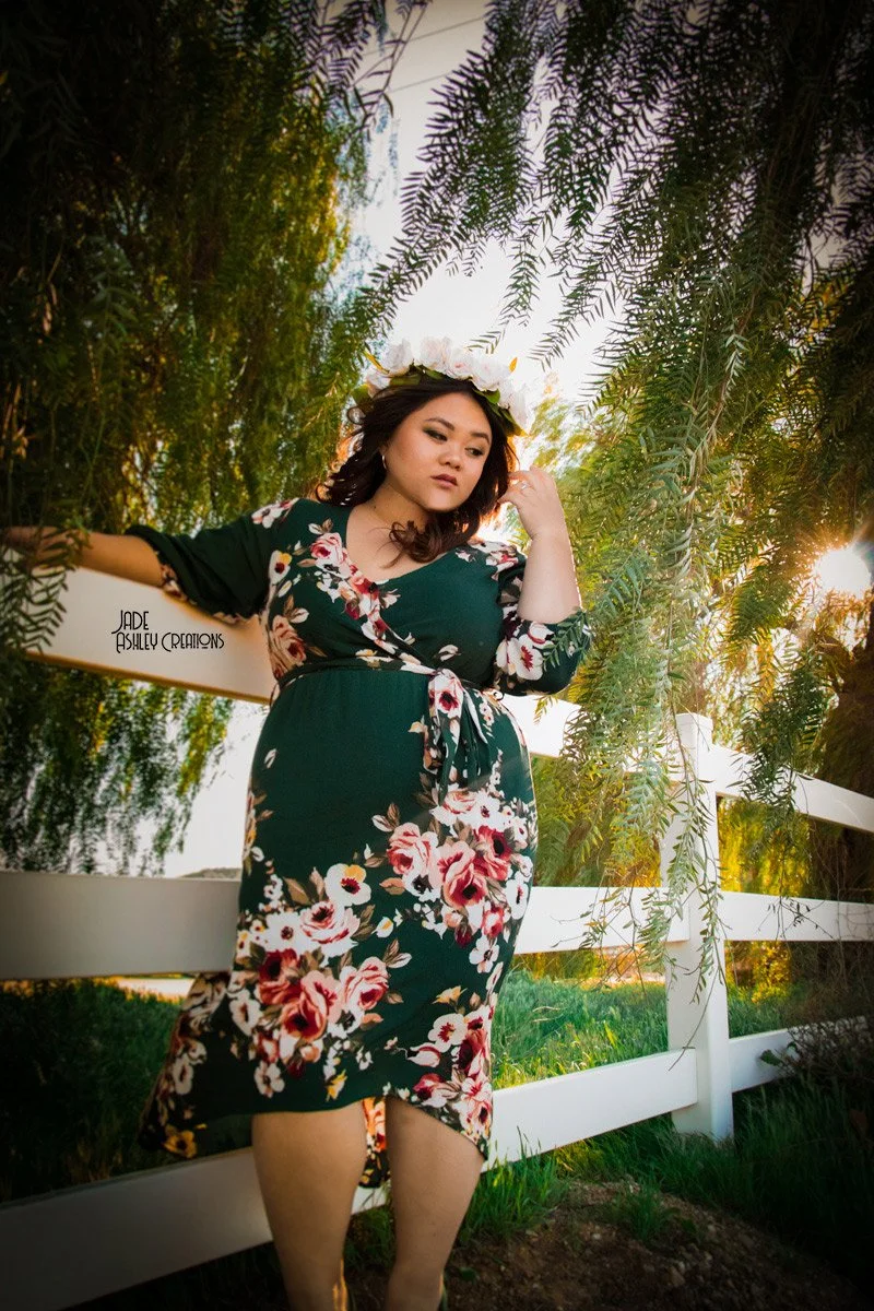 A woman with dark, shoulder-length hair and a floral dress standing outdoors next to a white fence and lush greenery, wearing a floral crown, with sunlight filtering through the trees.