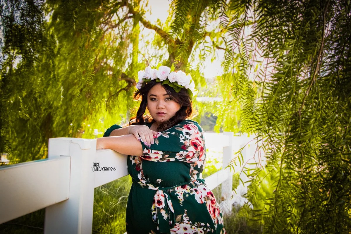 A woman with dark hair wearing a floral dress and a flower crown, leaning on a white fence in a lush green outdoor setting with sunlight filtering through trees.