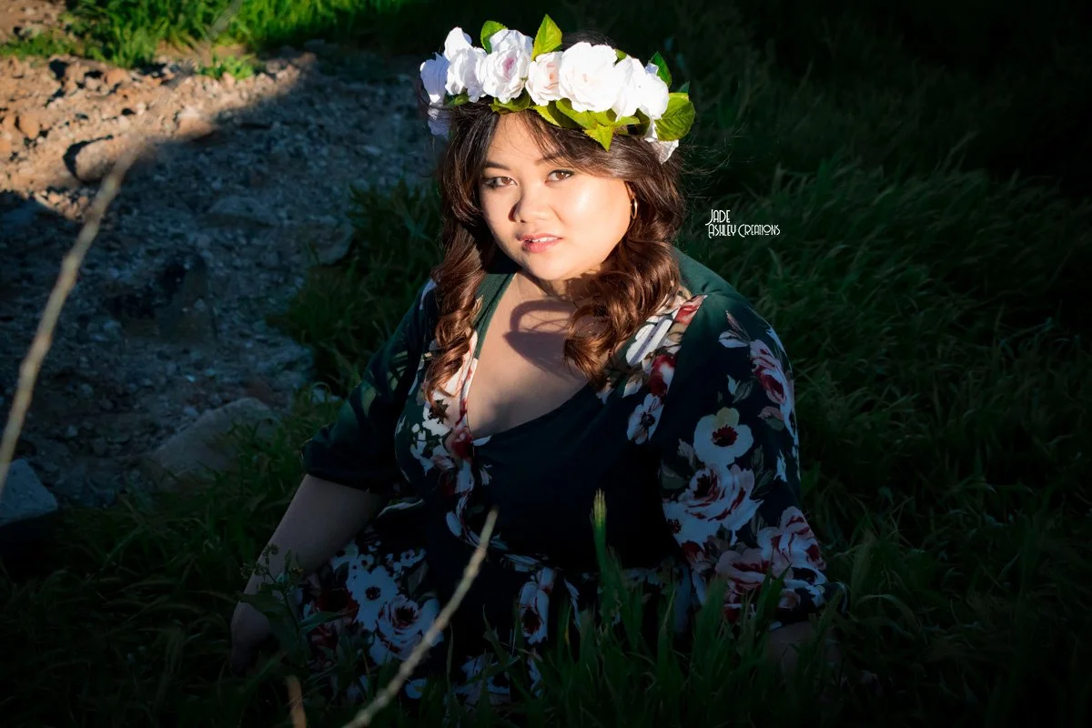 A woman with long brown hair wearing a flower crown and a floral dress, sitting outdoors in a patch of tall grass and dirt, with sunlight illuminating her face.