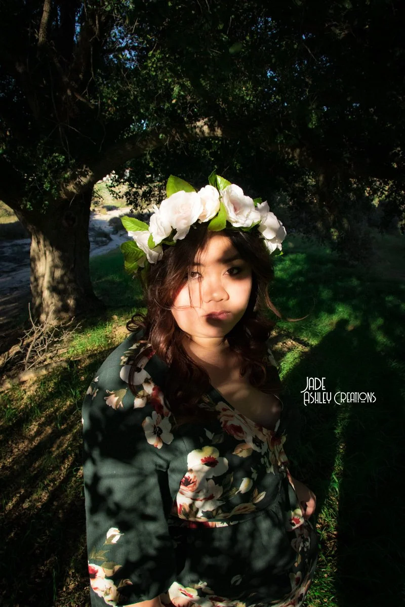 A woman standing outdoors under a large tree with sunlight and shadows on her face and clothing. She is wearing a floral dress and a white flower crown with green leaves.