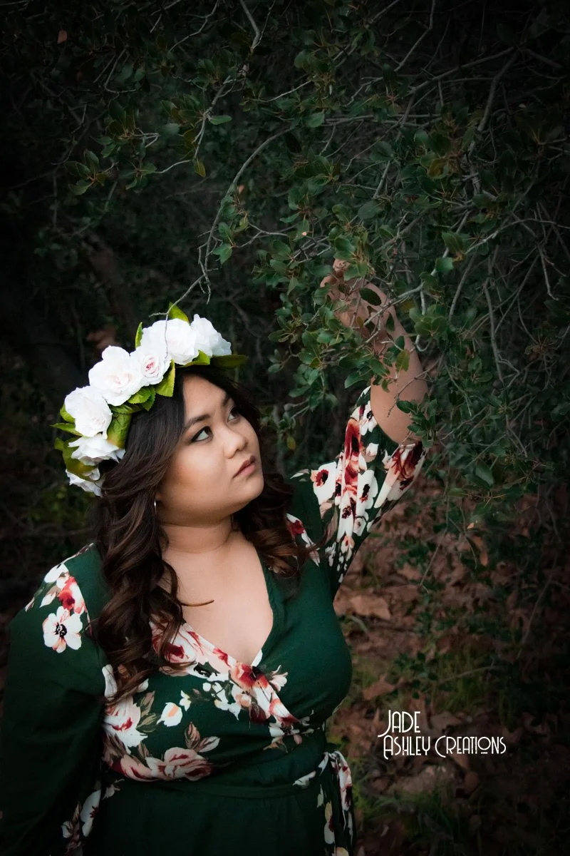 A woman wearing a green floral dress and a white flower crown is standing outdoors among tree branches, reaching up to touch the leaves.