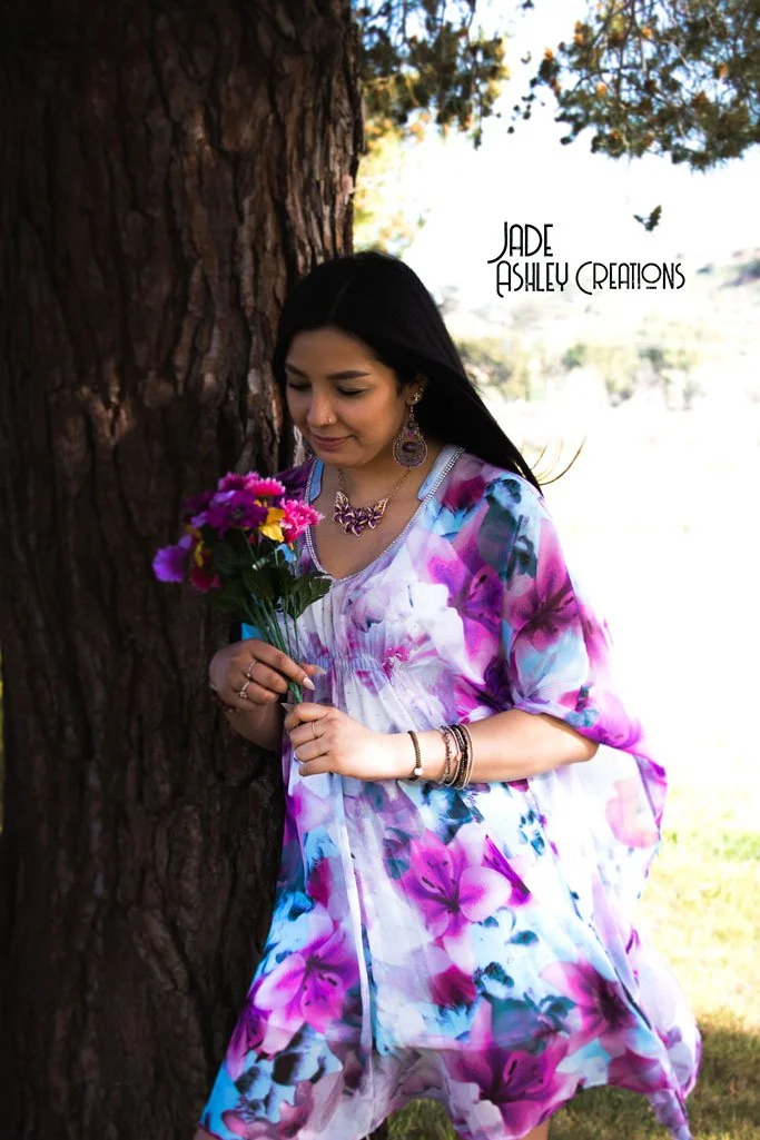 A woman with long dark hair wearing a colorful floral dress, jewelry, and a nose ring, holding a bouquet of purple and yellow flowers, standing next to a large tree outdoors.