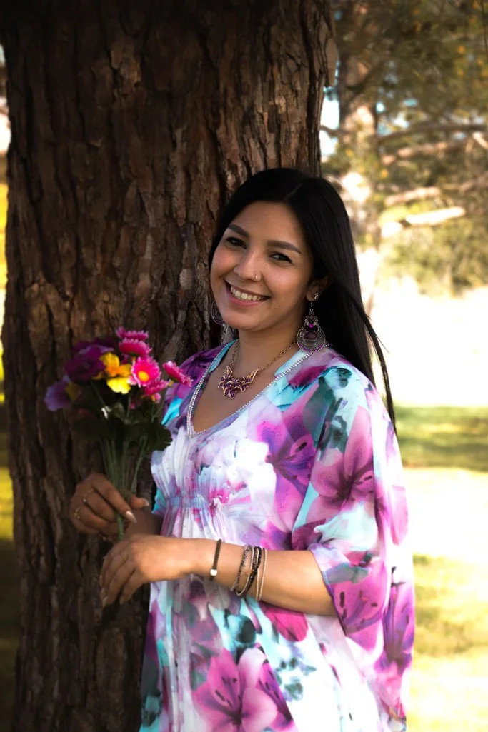 A woman with long dark hair smiling while holding a bouquet of colorful flowers outdoors near a large tree, wearing a vibrant floral dress and jewelry.