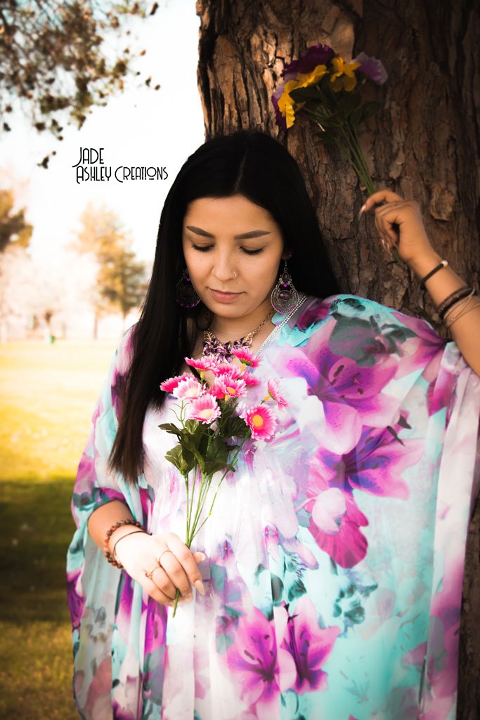 A woman with long black hair, wearing a colorful floral shawl, stands outdoors at a park, leaning against a tree with a small bouquet of pink flowers, holding a bunch of flowers in her hand, with yellow and purple flowers on the tree bark nearby.