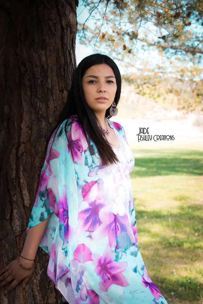 Young woman with long dark hair leaning against a tree outdoors, wearing a colorful floral dress and earrings, with a background of green grass and trees.