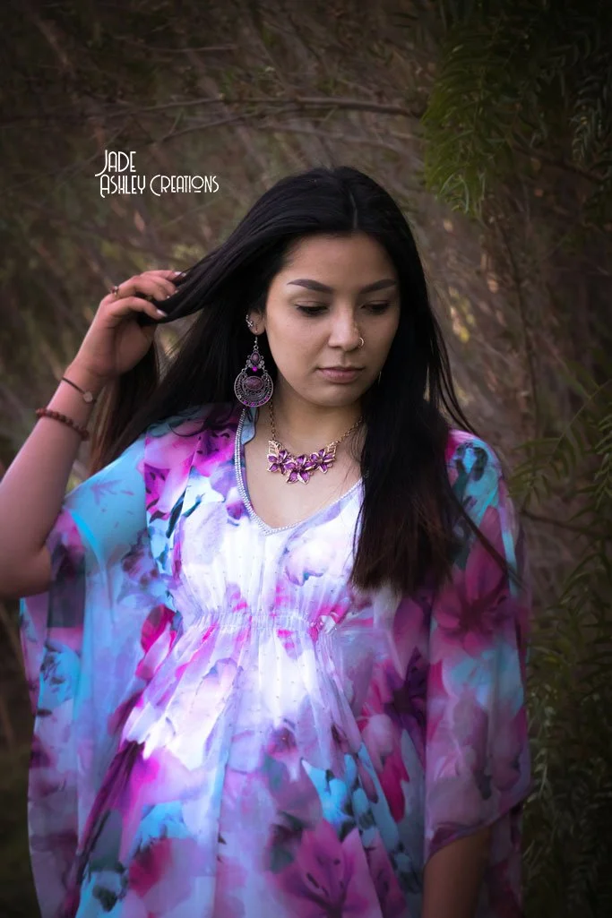 A young woman with long black hair, wearing a colorful, floral-patterned dress, and purple jewelry including earrings and a necklace, standing outdoors near a tree with dangling leaves.