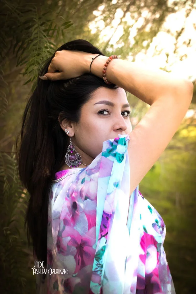 A woman with dark hair and earrings is posing outdoors with her right arm raised and hand resting on her head. She is wearing a colorful floral top, a beaded bracelet, and a black hair tie. The background includes greenery and sunlight filtering thro