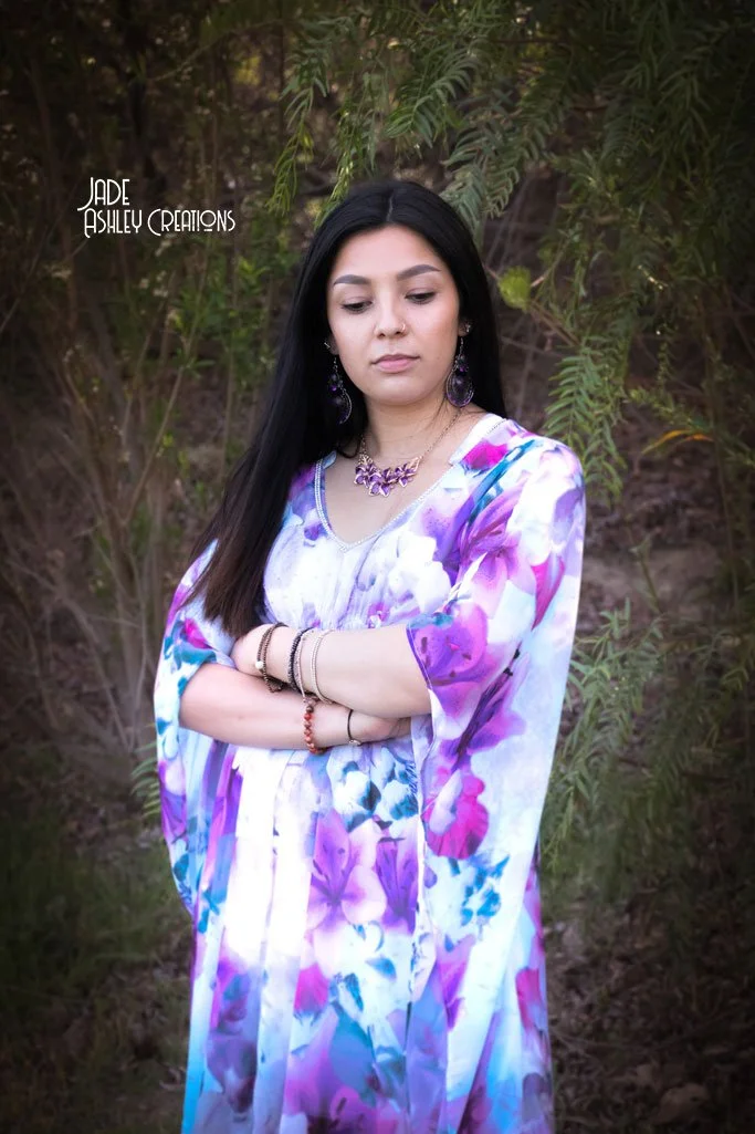 A woman with long black hair standing outdoors in front of green foliage, wearing a flowy floral dress with purple, pink, and blue flowers, accessorized with jewelry, with a serious expression and arms crossed.