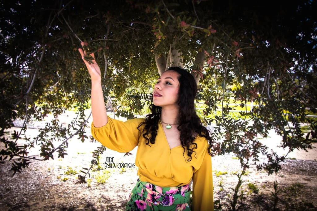 A woman with long dark curly hair wearing a yellow blouse and floral skirt standing under a tree, reaching towards the leaves, with sunlight filtering through the branches.