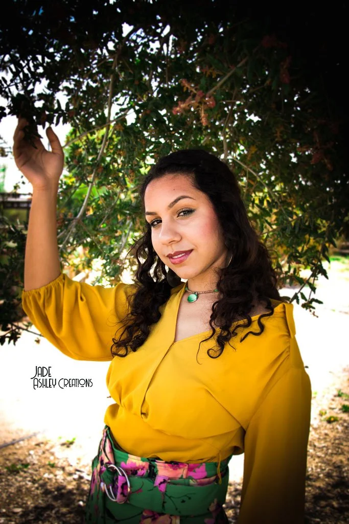 Young woman with curly dark hair, wearing a yellow blouse and green floral skirt, standing outdoors under a tree with leaves and branches, looking at the camera with a slight smile.