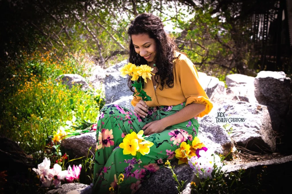 A woman sitting on rocks outdoors, holding yellow flowers, surrounded by greenery and colorful flowers, smiling and looking down.