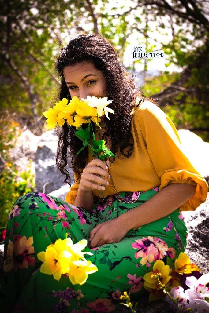 A woman with curly dark hair holding a bouquet of yellow flowers, sitting outdoors among greenery and rocks, wearing a yellow top and green floral pants.