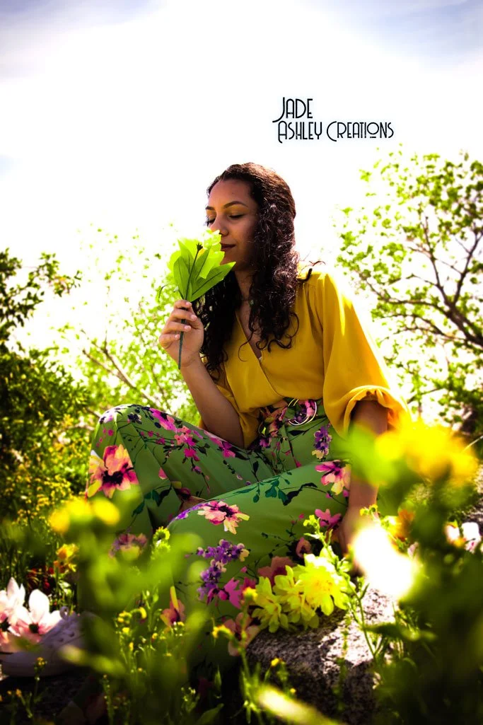 A woman with dark curly hair wearing a yellow top and floral green pants sitting outdoors among yellow and purple flowers, holding a green leaf close to her face with her eyes closed.