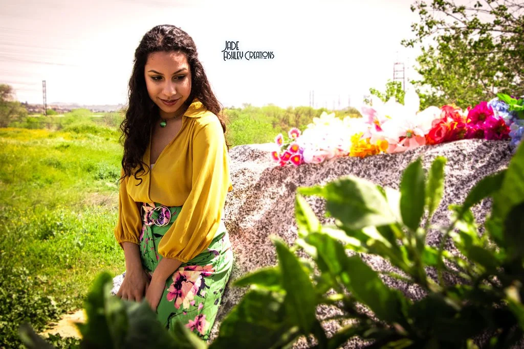 Woman in a yellow blouse and floral skirt standing outdoors near a large rock with colorful flowers on it, green grass and trees in the background.