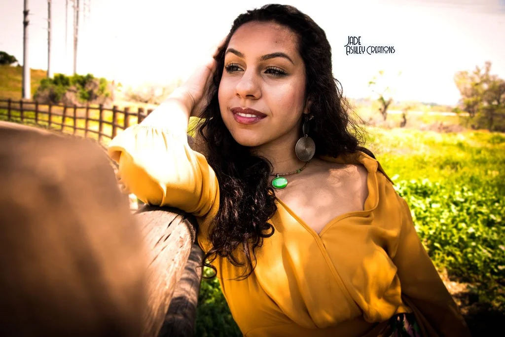 A young woman with dark, curly hair, wearing a yellow dress and large earrings, is leaning on a wooden fence outdoors, with a green field and trees in the background.