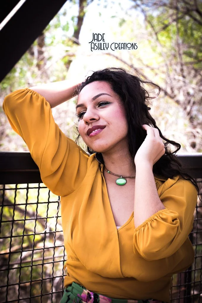 A woman with dark curly hair wearing a mustard yellow top and a green necklace, posing outdoors with a metal fence and trees in the background.