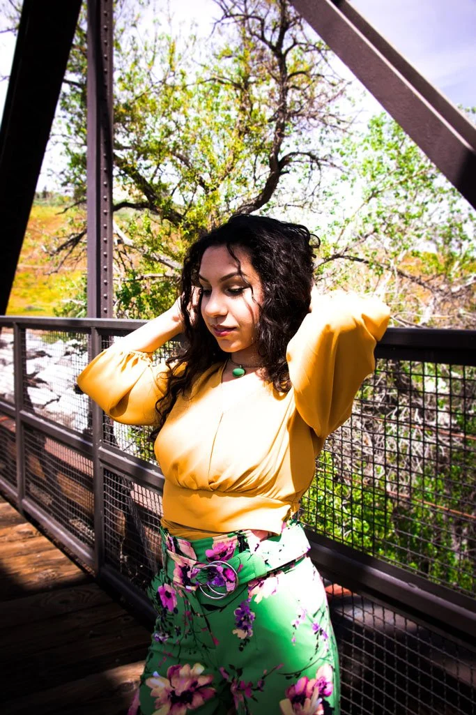 A woman with dark curly hair wearing a yellow top and green floral pants standing on a metal bridge with trees in the background.