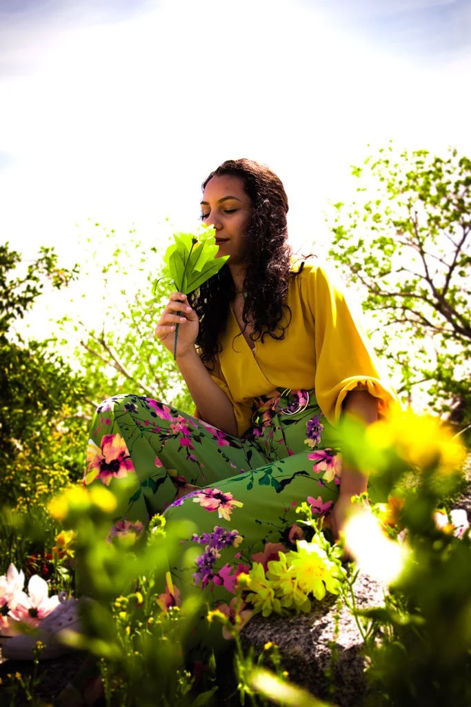 A woman in a yellow top and floral green pants sitting outdoors among colorful flowers, holding a green leaf close to her face, surrounded by lush greenery and trees in the background.