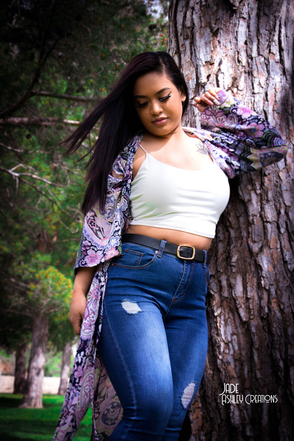 A young woman with long dark hair leaning against a tree outdoors, wearing a white crop top, ripped blue jeans, and a colorful floral jacket.
