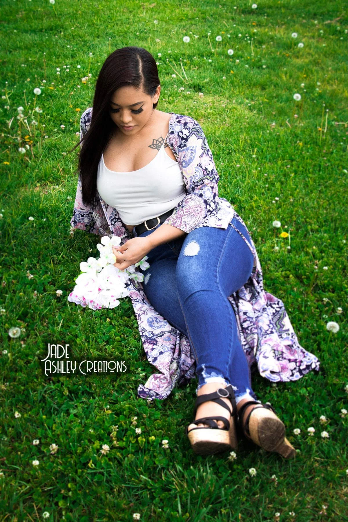 A woman sitting on a grassy field holding white flowers. She has long dark hair, a tattoo on her shoulder, and is wearing a white top, a patterned long cardigan, ripped jeans, and sandals.