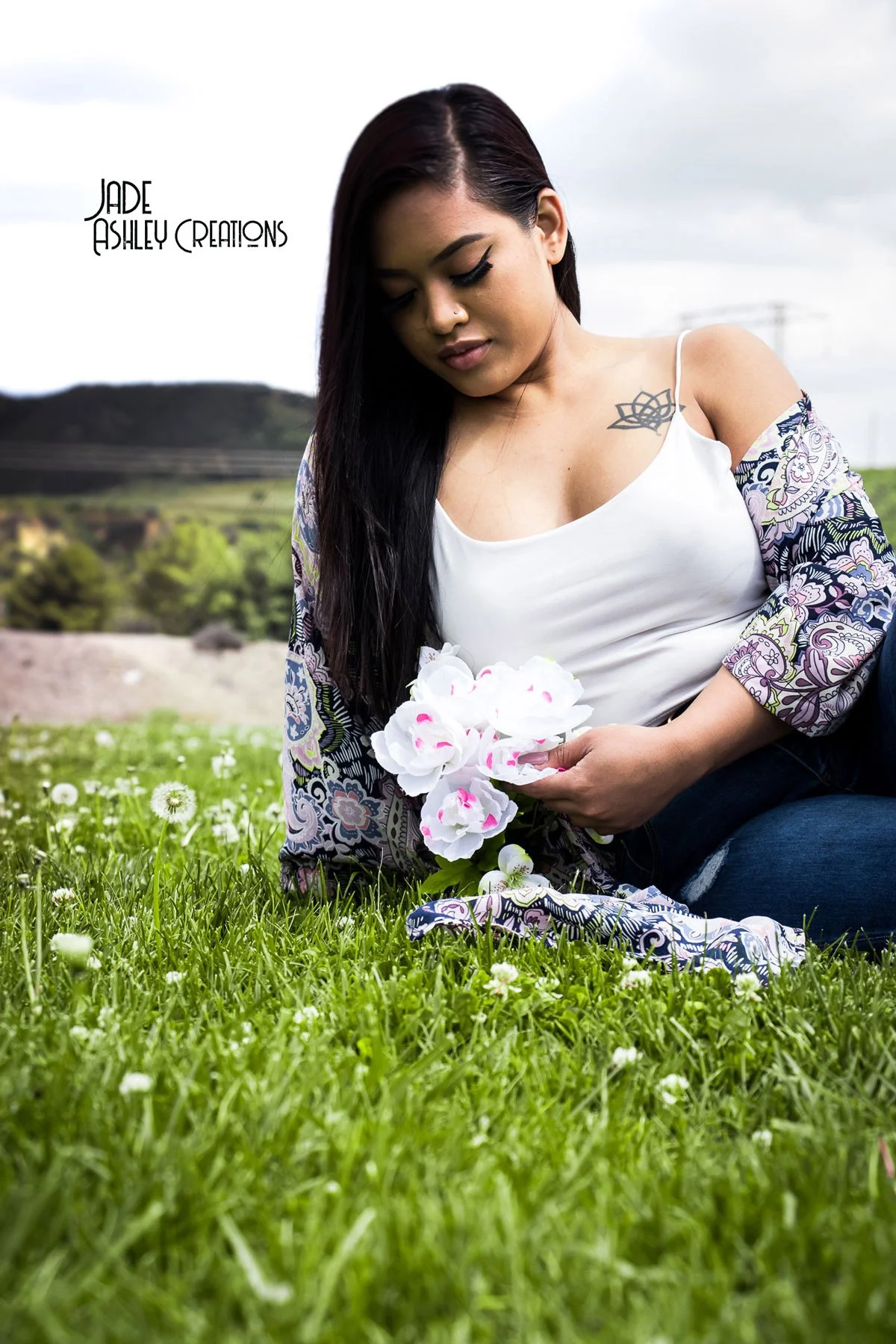 A woman sitting on grass in a field holding white flowers, with a blurred background of trees and hills.