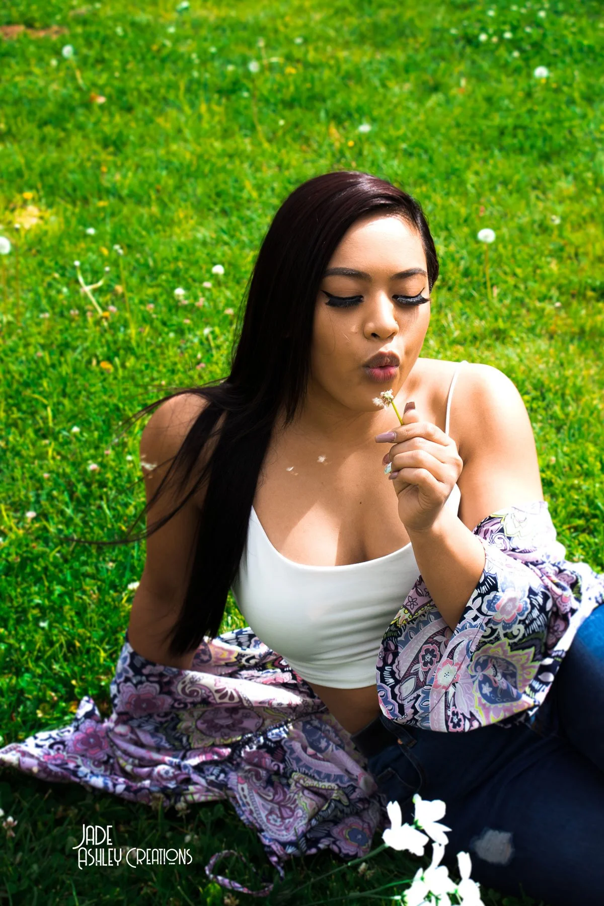 Young woman with long dark hair sitting on lush green grass surrounded by small white flowers, holding a small flower near her lips, with her eyes closed and a peaceful expression.