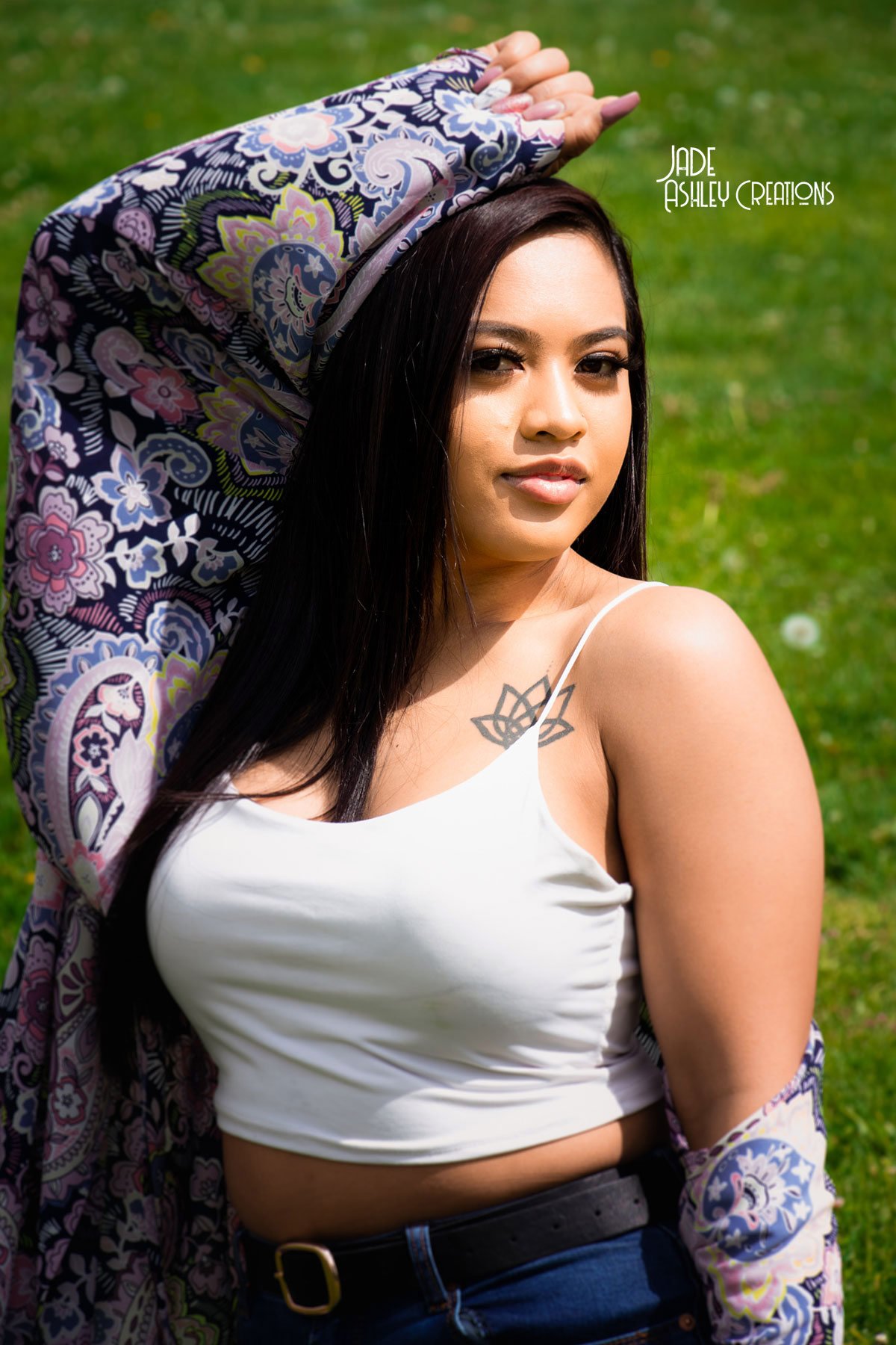A woman with long dark hair wearing a white tank top, dark jeans, and a black belt, standing outdoors on grass, with a colorful paisley-patterned shawl over her head and shoulders. She has a tattoo of a lotus flower on her shoulder and is posing with