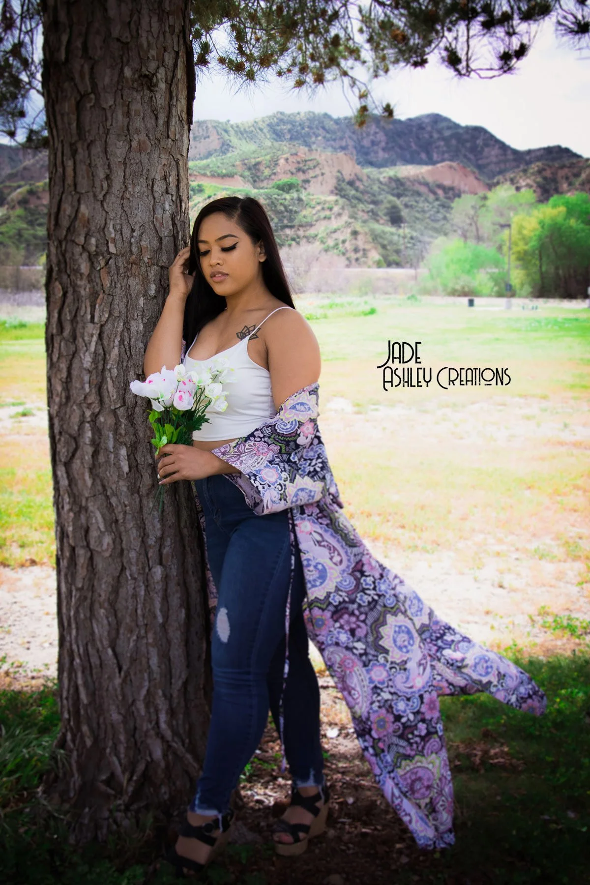 A young woman standing by a tree in a scenic outdoor setting with mountains and green trees in the background, holding a bouquet of white and pink flowers, wearing a white tank top, dark jeans, and a colorful kimono-style cover-up.