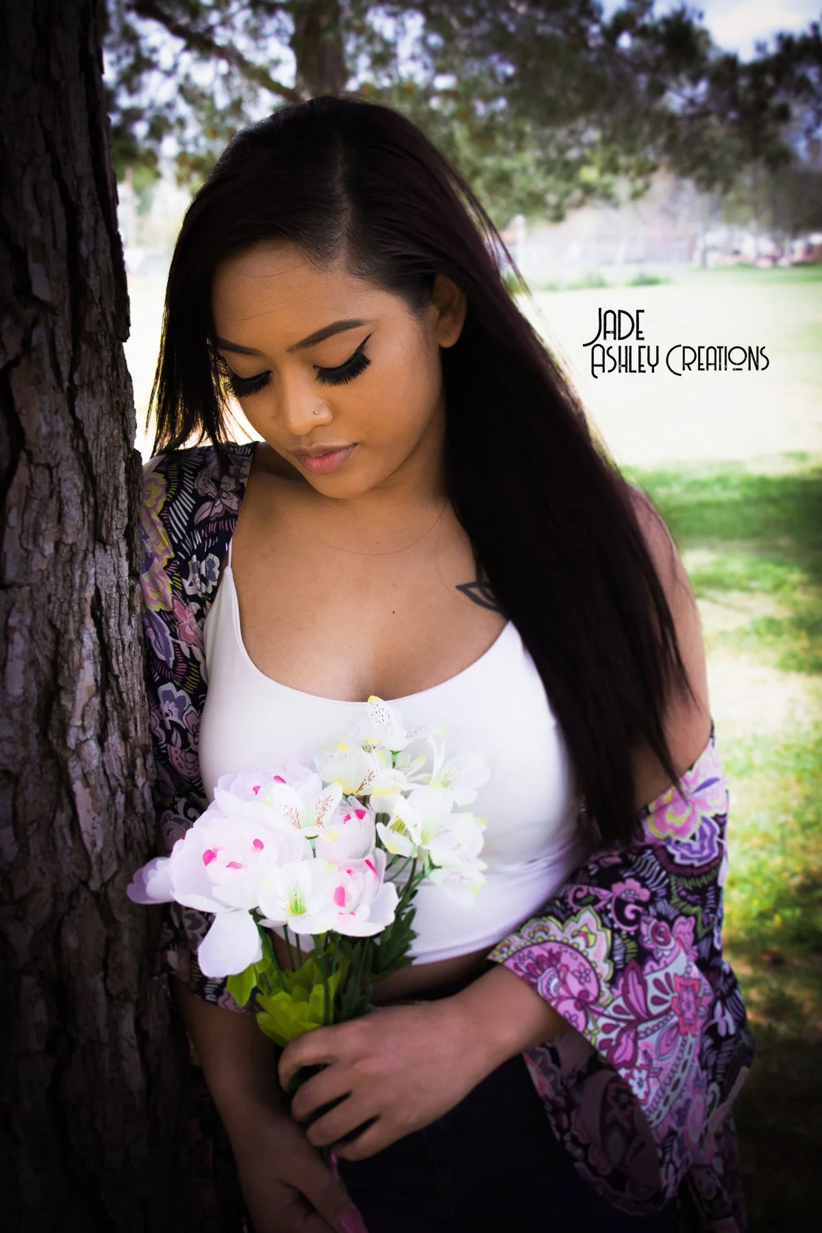 A woman with dark hair and makeup, holding a bouquet of white and pink flowers, standing outdoors near a tree in a park.
