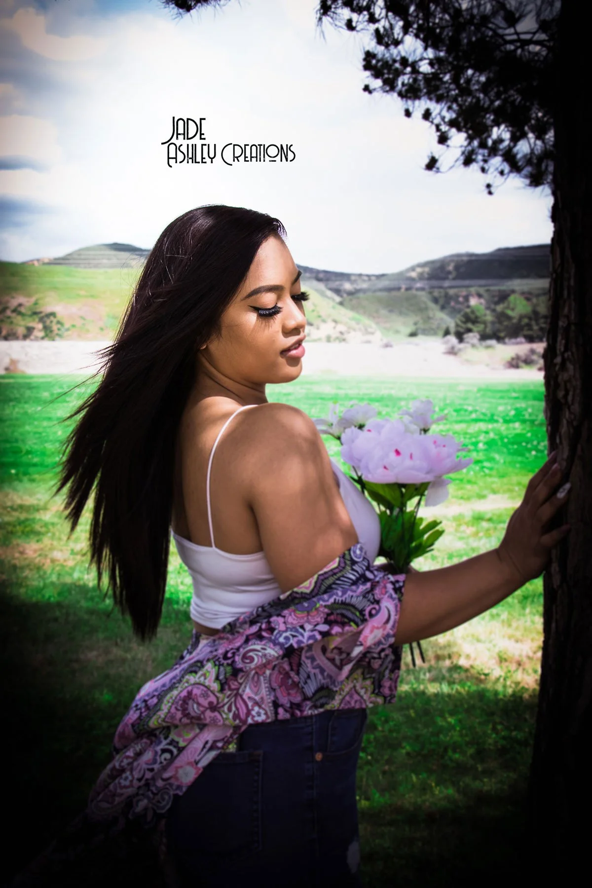 A young woman with long dark hair standing near a tree, holding a bouquet of white flowers, with rolling green hills and a partly cloudy sky in the background.