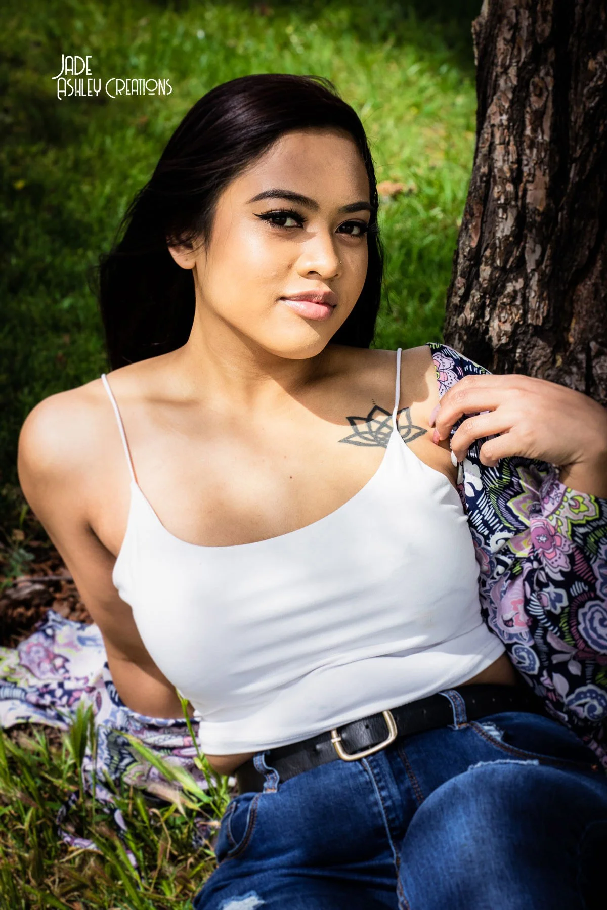A woman with long dark hair, sitting outdoors, leaning against a tree. She is wearing a white tank top, blue jeans, and has a tattoo near her collarbone. The background includes green grass.