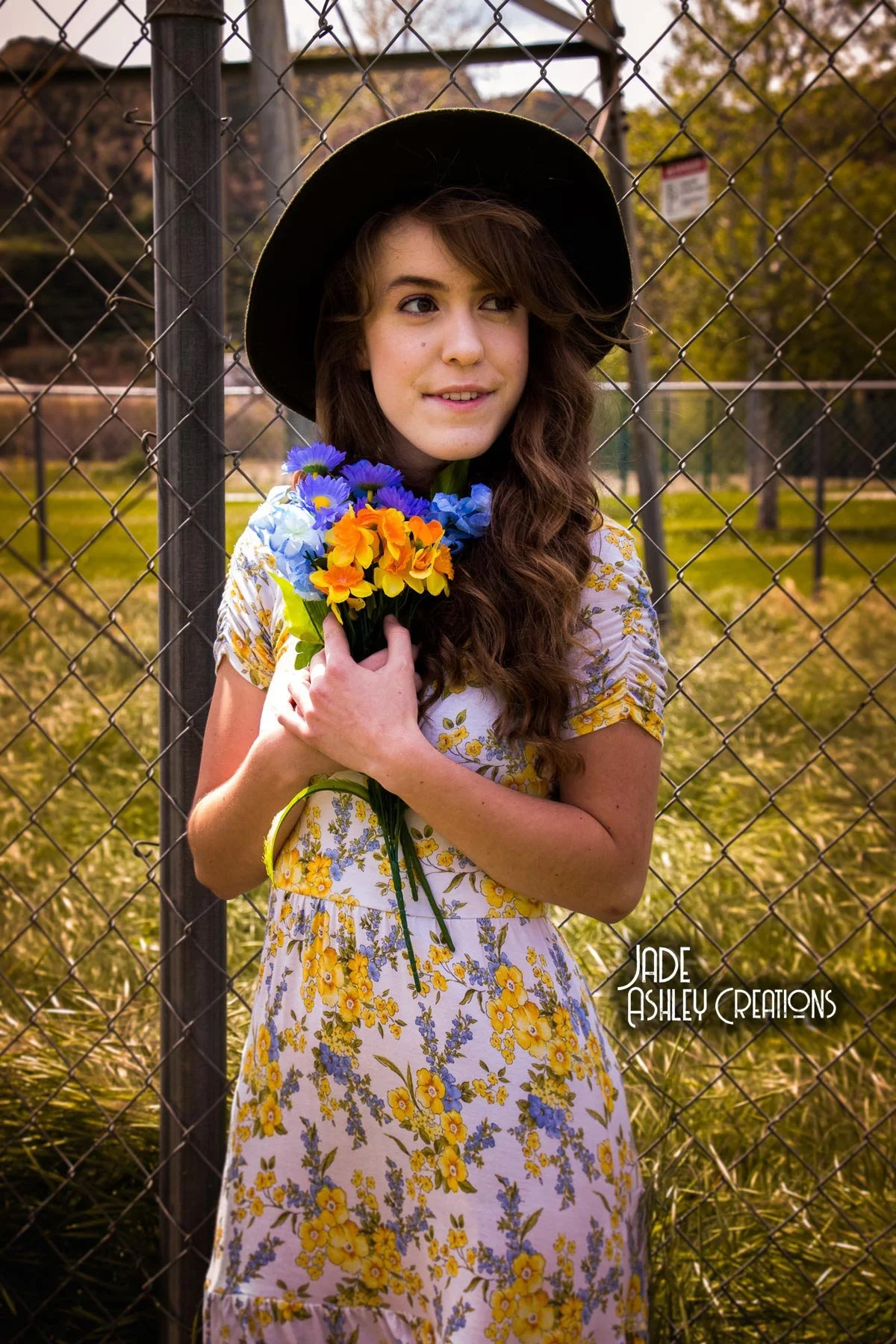 A young woman with long, wavy brown hair wearing a large black hat and a yellow floral dress, holding a bouquet of blue, orange, and yellow flowers, standing in front of a chain-link fence in a park with autumn trees in the background.