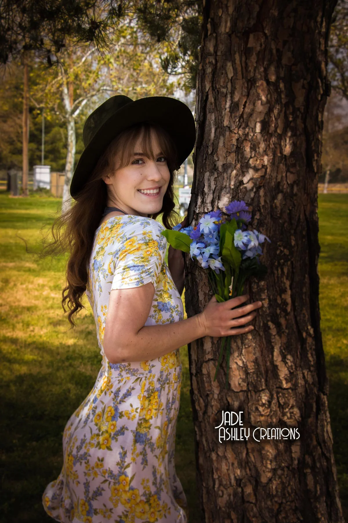 A smiling woman with long brown hair, wearing a wide-brimmed black hat and a floral dress with yellow and blue flowers, stands next to a tree holding a bouquet of blue and purple flowers. She is outdoors in a park with green grass and trees in the ba