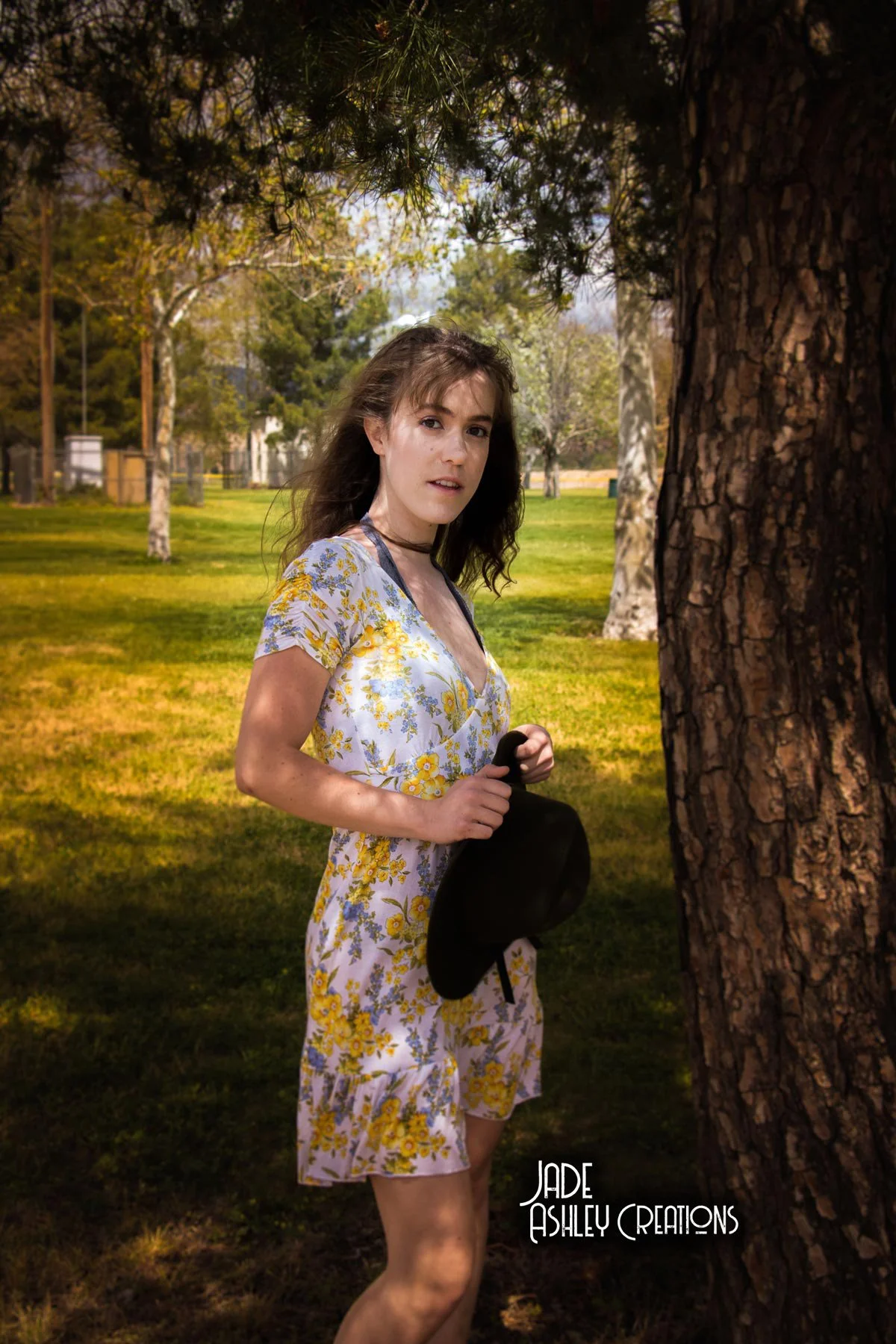 A young woman wearing a floral dress holding a black hat stands behind a tree in a park filled with trees and grass.