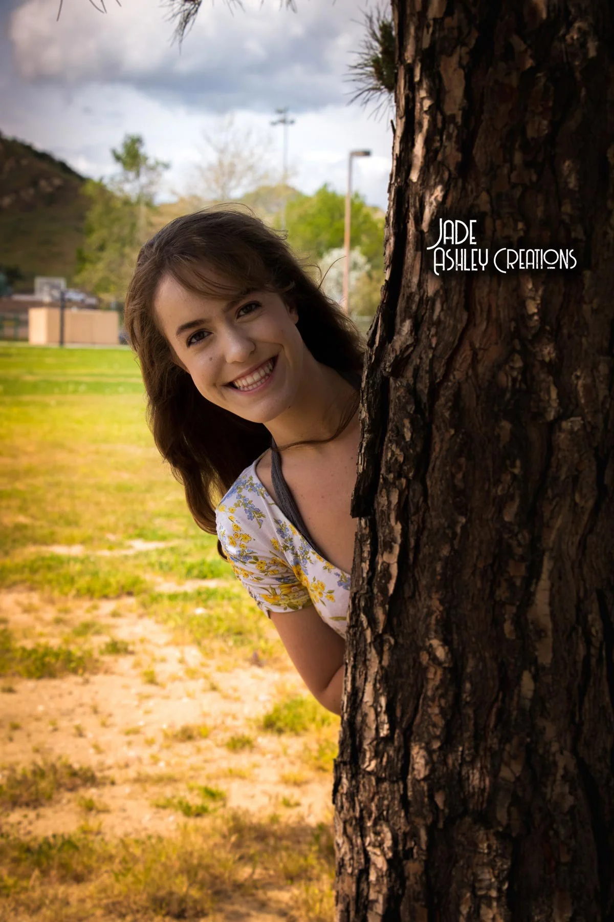 A young woman peeking from behind a tree with a big smile in a park. She has brown hair and is wearing a floral top. There are trees, a grassy area, and cloudy sky in the background.