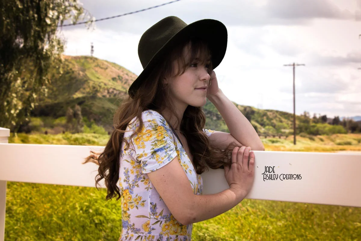 A young woman with long brown hair and freckles wearing a wide-brimmed black hat and a white floral dress, resting her arms on a white fence in a rural landscape with green hills and a cloudy sky.