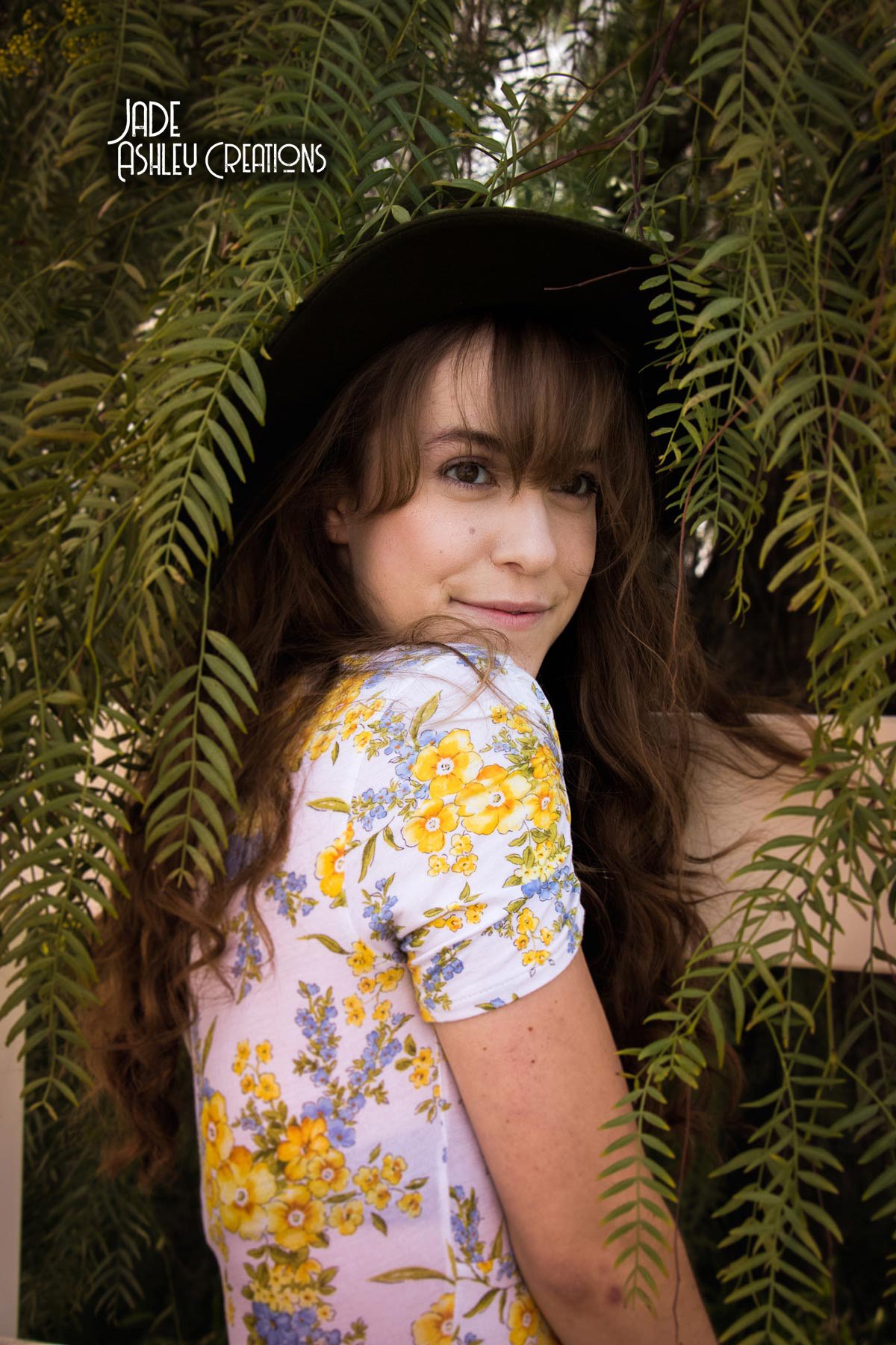 A young woman with long brown hair and blue eyes, smiling softly, is sitting among green ferns. She is wearing a wide-brimmed black hat and a white dress with yellow and blue floral print.