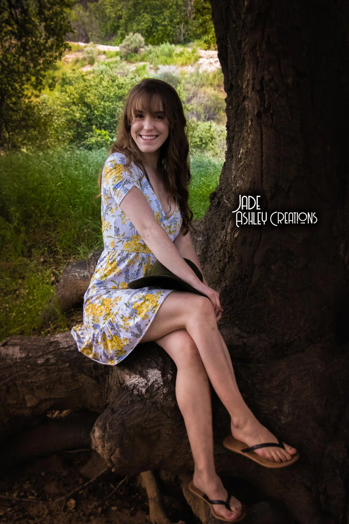 A woman with long brown hair and a big smile, wearing a floral dress and flip-flops, sitting on a tree root in a lush green outdoor setting.