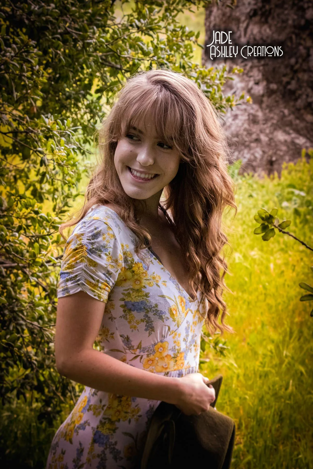 A woman with long, wavy red hair smiling outdoors, wearing a floral dress with yellow and purple flowers, surrounded by green foliage and trees.