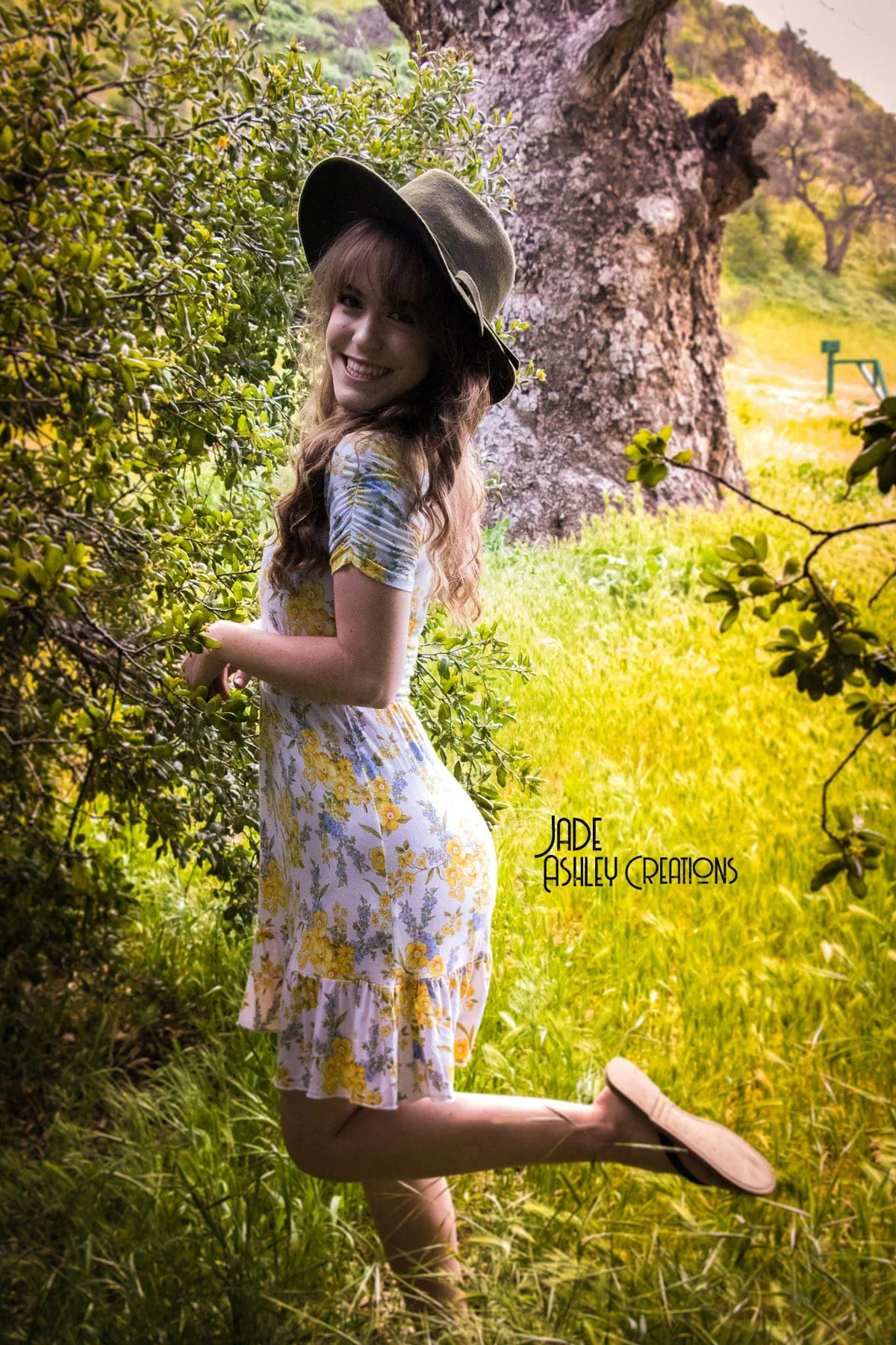Young woman smiling and posing outdoors in a grassy area with trees and large tree trunk in the background, wearing a floral dress and a wide-brimmed hat.