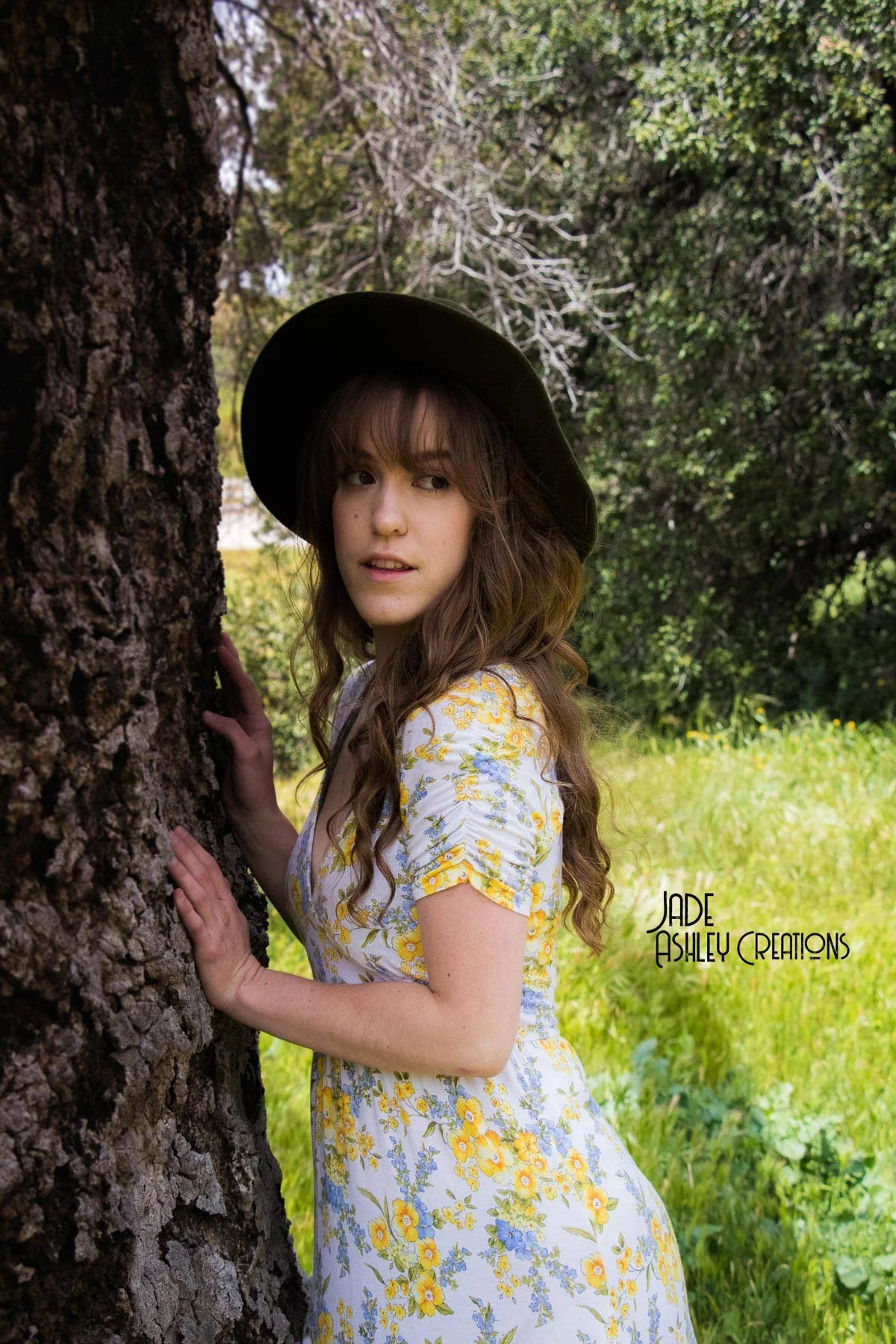 A woman with wavy brown hair wearing a black wide-brimmed hat and a floral dress with yellow and blue flowers, standing outdoors behind a tree in a green grassy area.