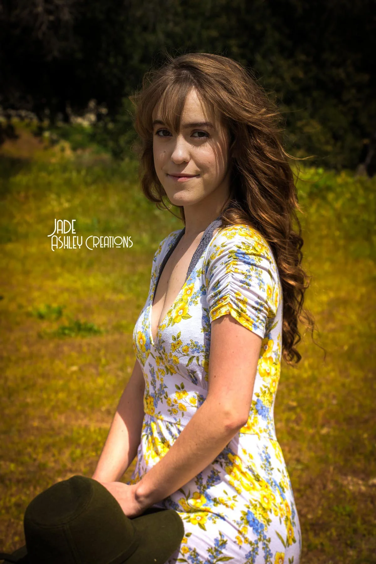 Young woman with long, curly brown hair wearing a yellow and blue floral dress. She is standing outdoors in a grassy area with trees in the background, holding a black hat.