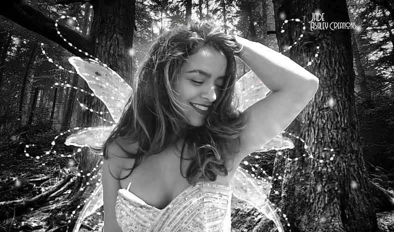 Black and white photo of a woman with fairy wings smiling in a forest, wearing a strapless dress, with her hand in her hair.